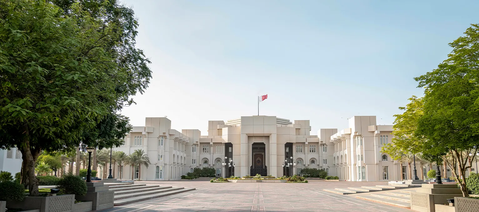 The white stone facade of the Amiri Diwan in Doha, Qatar, with the Qatari flag on the roof, seen across a paved forecourt framed by trees under a clear sky