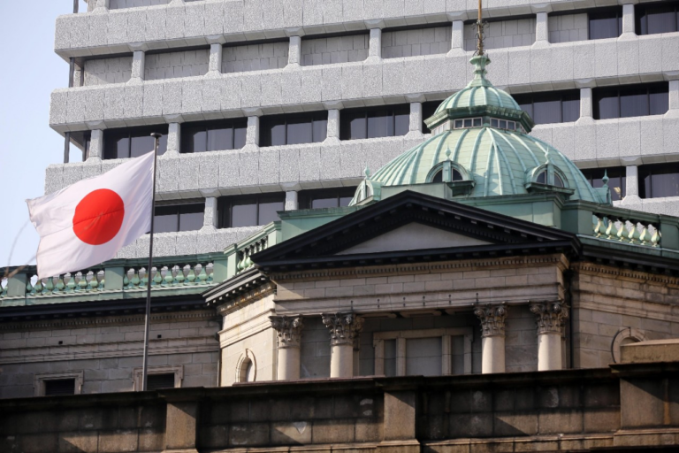 The Bank of Japan head office in Tokyo, with its green copper dome and stone columns in front of a modern office tower, and the Japanese flag flying on the left.