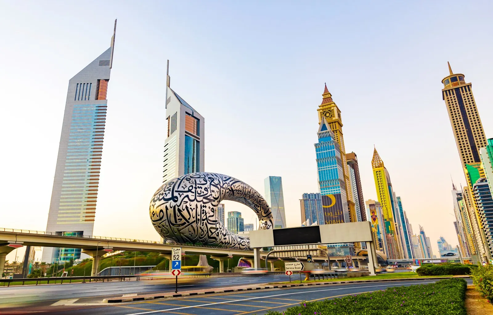 The Museum of the Future, its steel facade wrapped in Arabic calligraphy, beside the twin Emirates Towers and the high-rises of Sheikh Zayed Road in Dubai, with traffic on the highway in the foreground