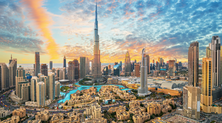 Aerial view of downtown Dubai at sunset, with the Burj Khalifa rising above surrounding towers, the Dubai Fountain lake and low-rise buildings in the foreground.