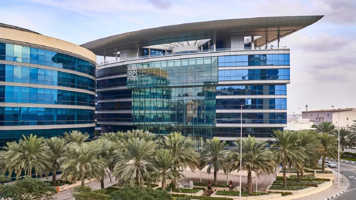The glass and steel Dubai Airport Freezone building, with its name on the facade and a curved roof canopy, behind rows of date palms under a cloudy sky