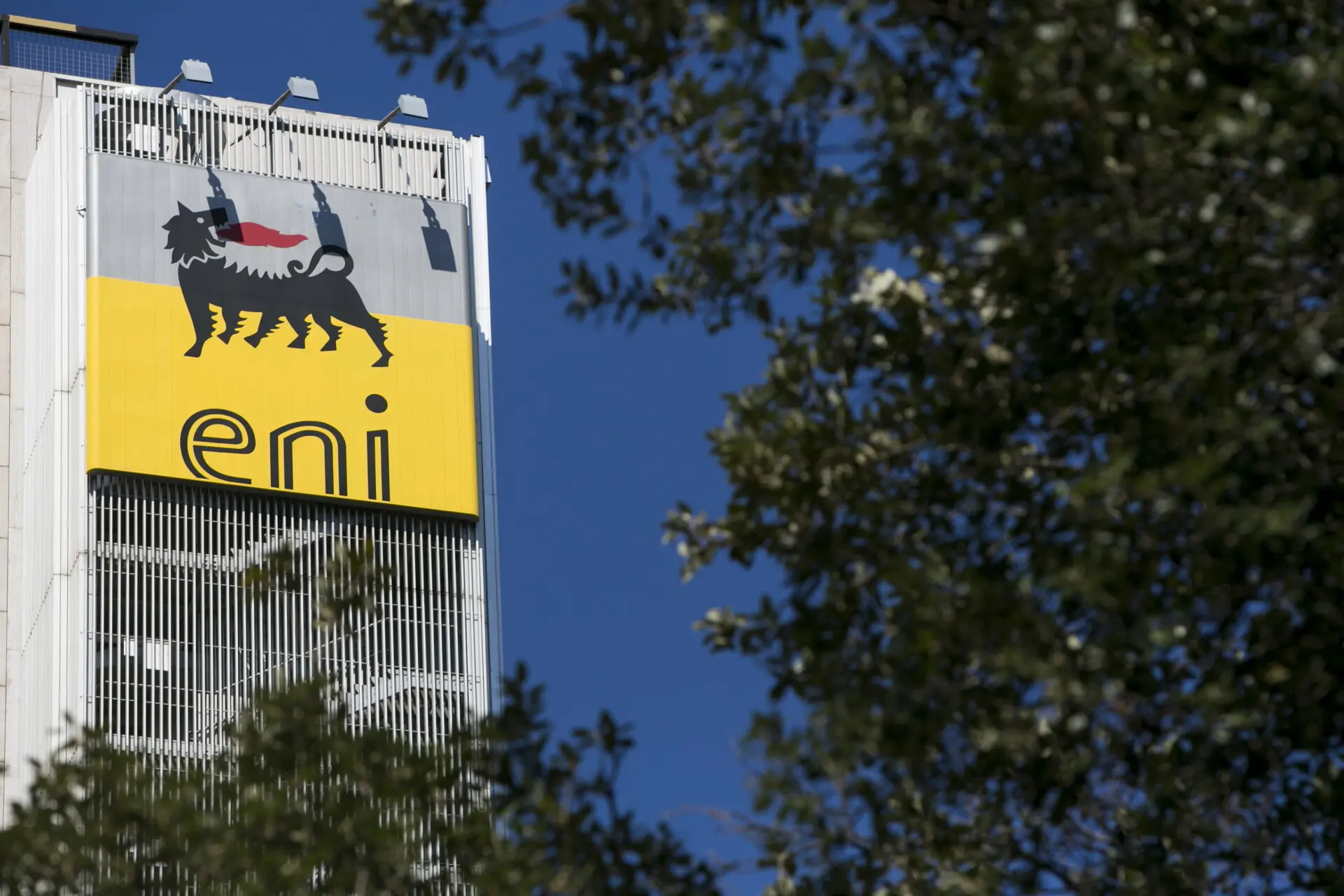 An office tower bearing the Eni logo, the six-legged dog above the name eni on a yellow panel, against a blue sky with trees in the foreground