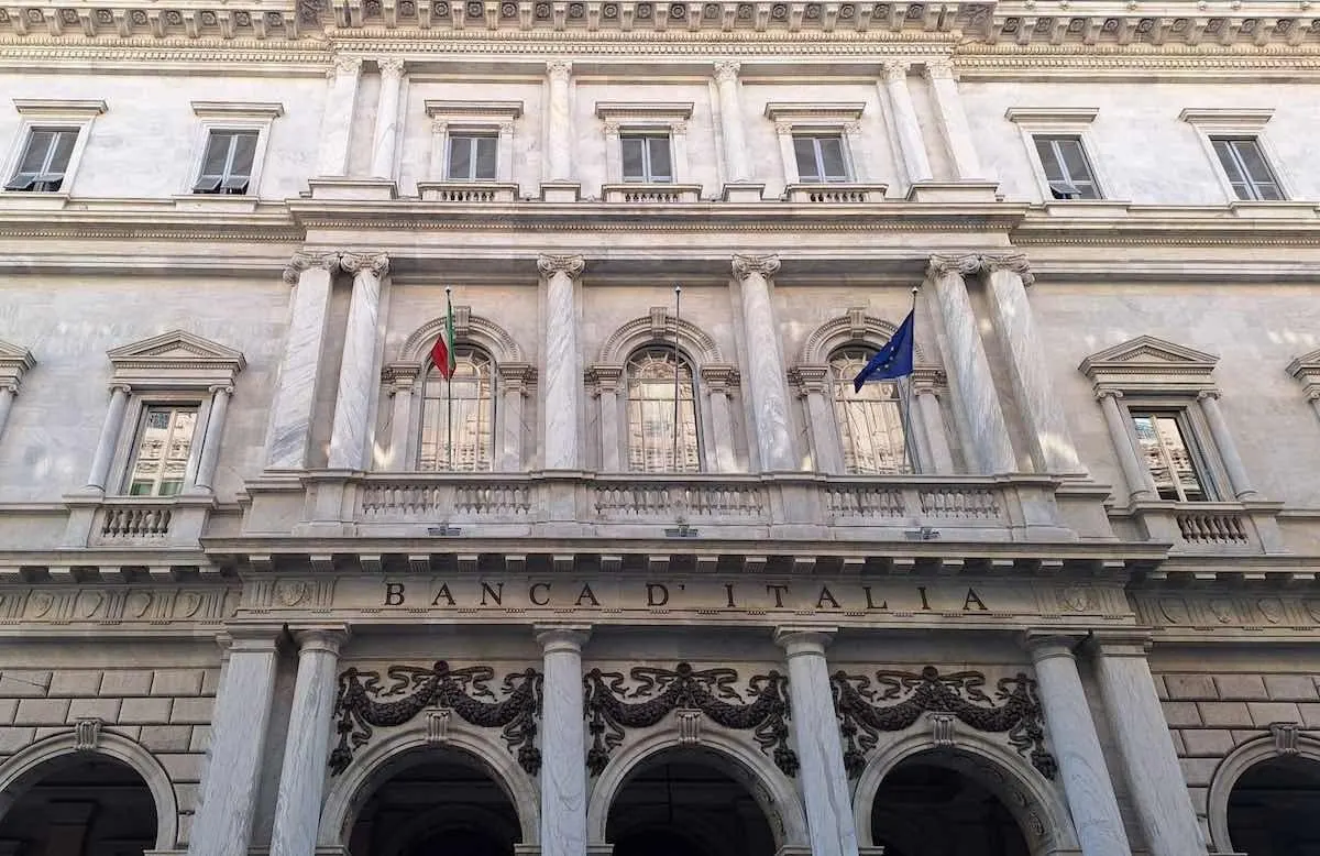 The stone facade of a Banca d'Italia building, with the bank's name carved above arched entrances, columns and balconies, and the Italian and European Union flags