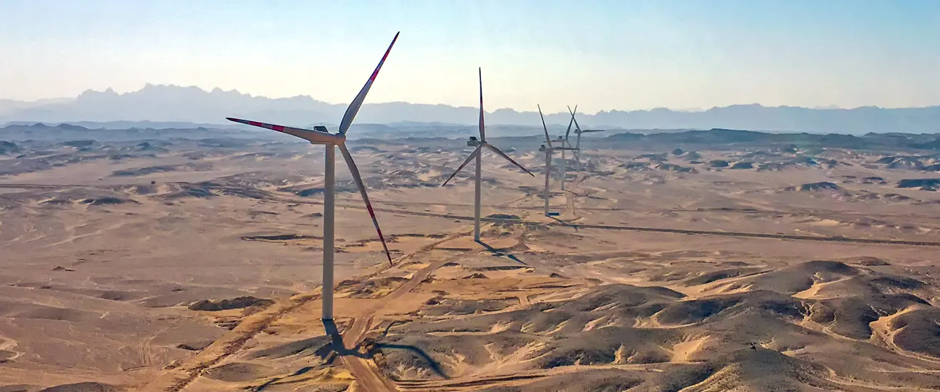 Wind turbines standing on desert ground at a Gulf of Suez wind farm in Egypt, with mountains on the horizon