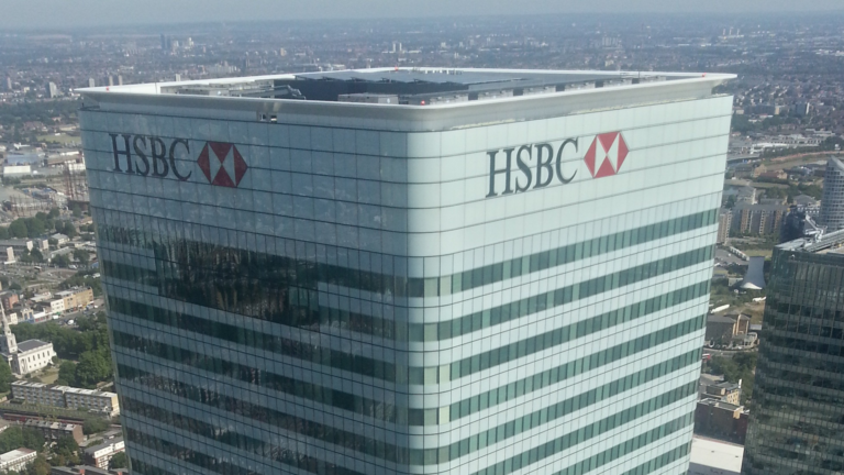 The red and white HSBC logo on the top of the HSBC tower in London, seen from above with the city behind