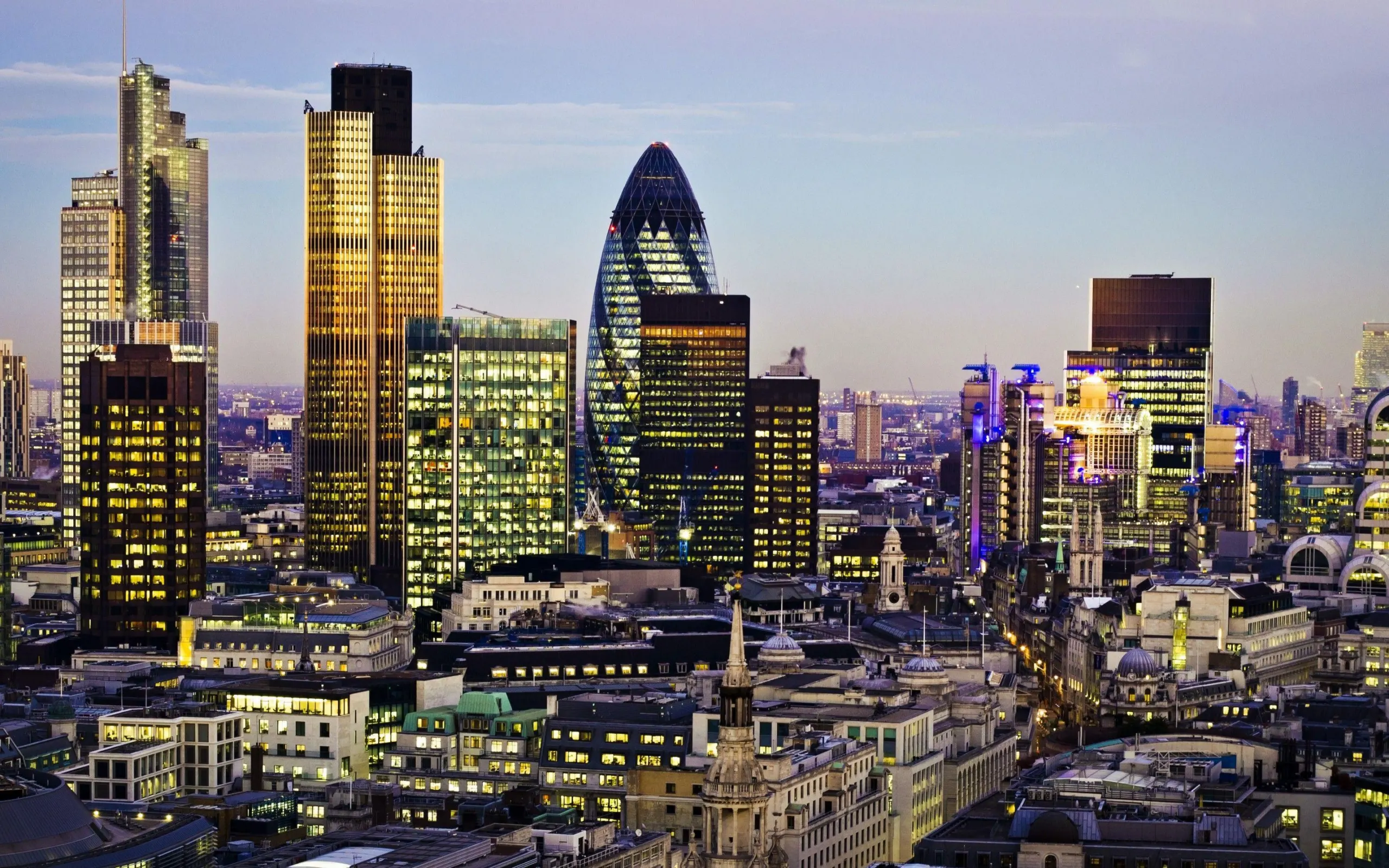 The City of London financial district skyline at dusk, with the curved glass 30 St Mary Axe tower, known as the Gherkin, among lit office towers above older stone buildings and church spires