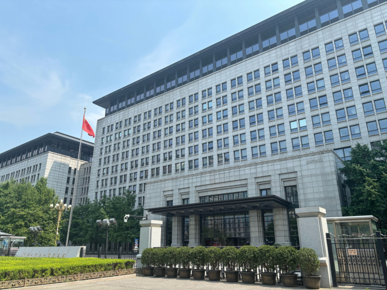 A large government office building with a Chinese flag on a flagpole and a gated entrance with hedges