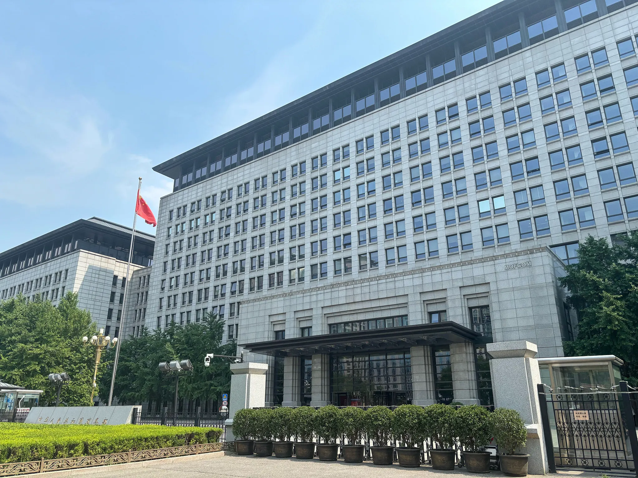 A large government office building with a Chinese flag on a flagpole and a gated entrance with hedges