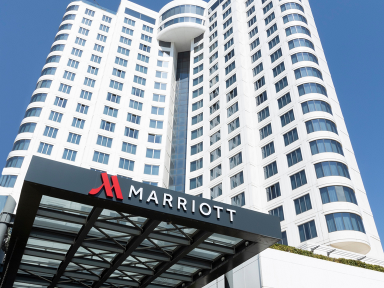 A white Marriott hotel tower under a clear blue sky, with the red Marriott logo and name on the entrance canopy