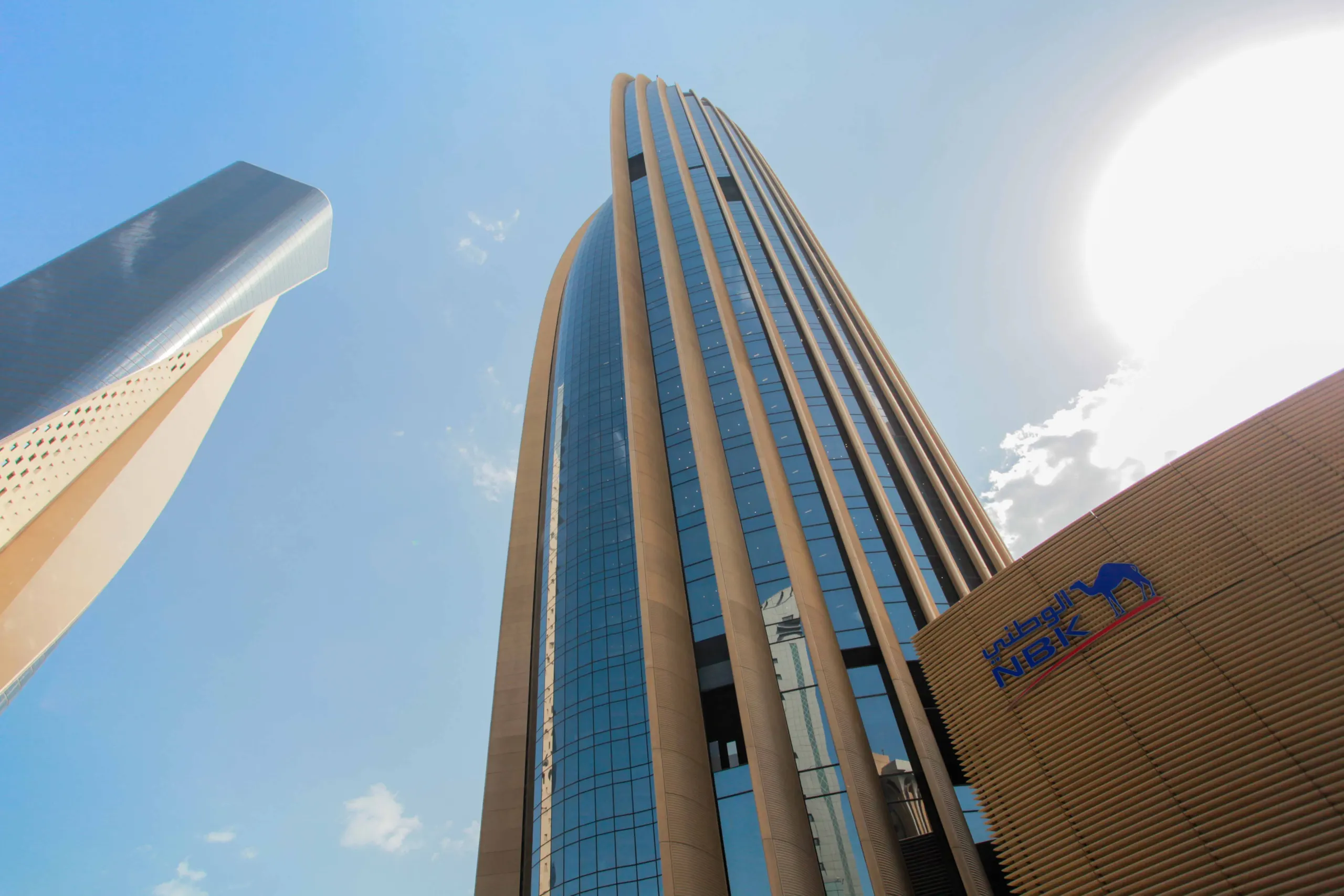 The National Bank of Kuwait headquarters tower, its curved glass facade framed by vertical fins, with the NBK logo on the building in front, seen from street level against a blue sky