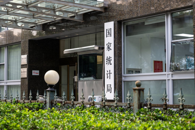 The entrance of the National Bureau of Statistics of China, its name in Chinese characters on a tall white sign beside a glass canopy, behind an iron fence and a green hedge