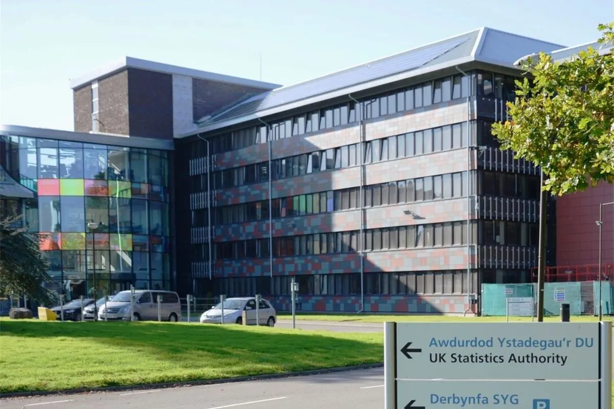 An Office for National Statistics building with a glass and patterned panel facade, behind a bilingual Welsh and English sign pointing to the UK Statistics Authority and ONS reception