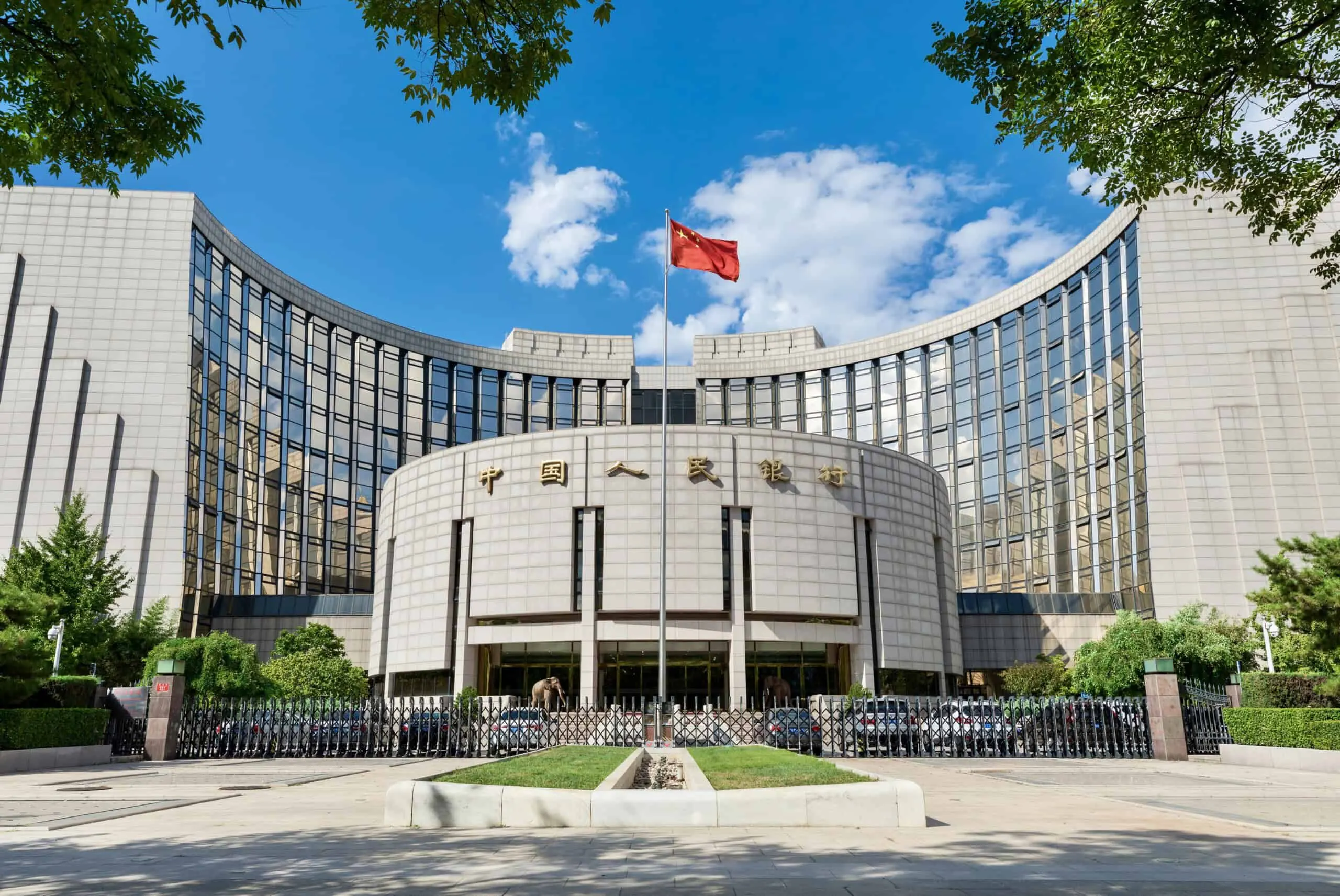 The curved stone and glass headquarters of the People's Bank of China, with the bank's name in gold Chinese characters above the entrance and the Chinese flag flying in front, under a blue sky