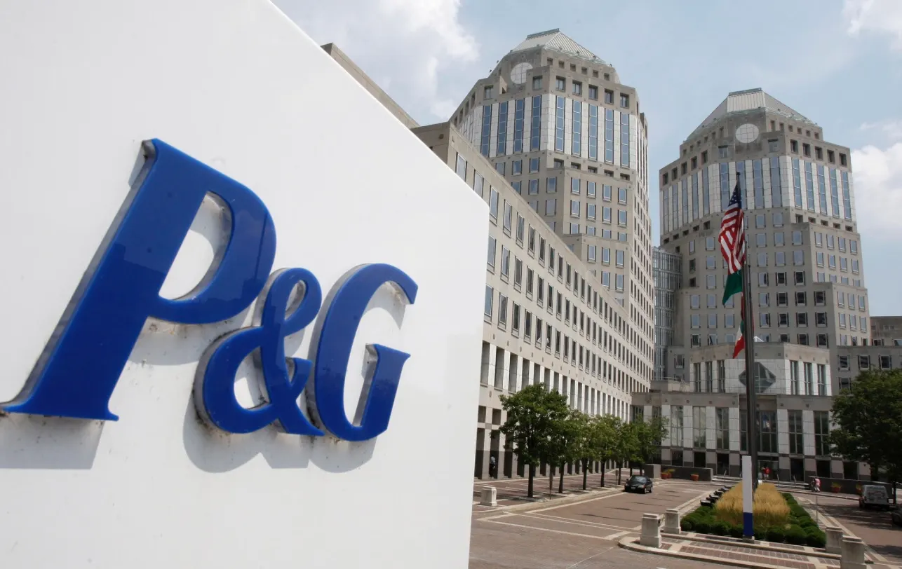 A blue P&G logo sign in front of the company's twin-towered headquarters, with flags on a flagpole