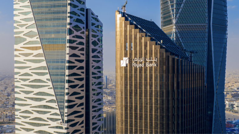 A bronze-toned office tower carrying the Riyad Bank logo and its Arabic and English names, between two modern towers