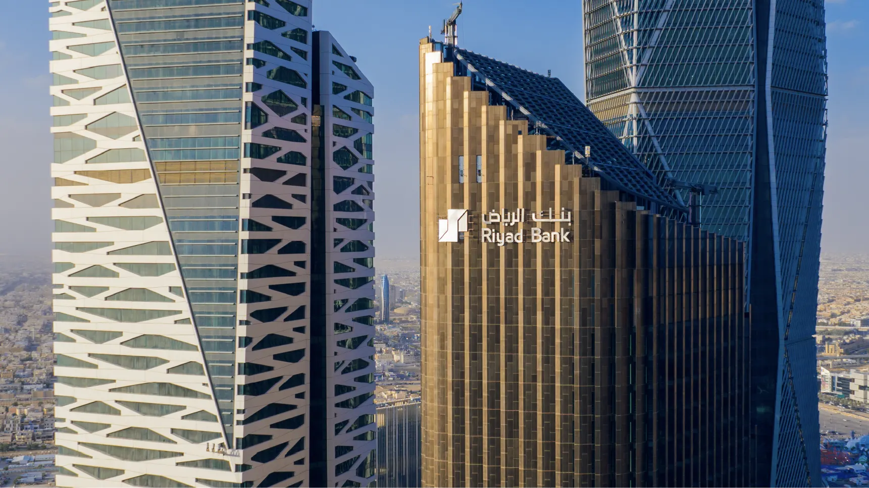 A bronze-toned office tower carrying the Riyad Bank logo and its Arabic and English names, between two modern towers