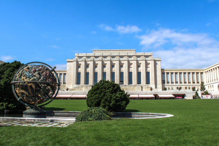 The Palais des Nations in Geneva, home of UNCTAD, seen across a green lawn with the bronze celestial sphere in the foreground under a blue sky