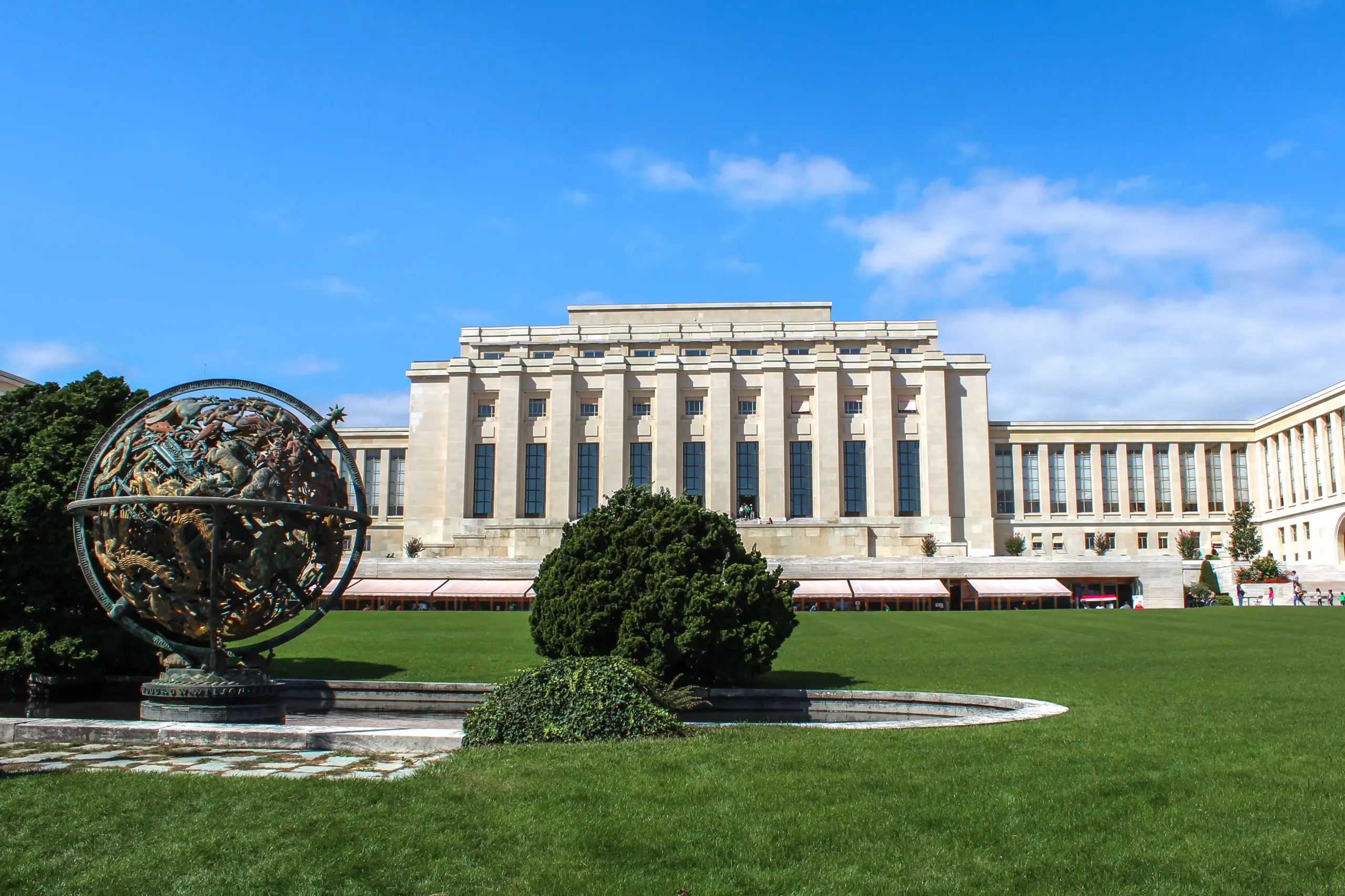 The Palais des Nations in Geneva, home of UNCTAD, seen across a green lawn with the bronze celestial sphere in the foreground under a blue sky