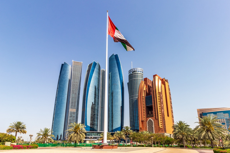 The UAE flag flying on a tall flagpole in Abu Dhabi in front of the Etihad Towers and other high-rise buildings, with palm trees under a clear blue sky.