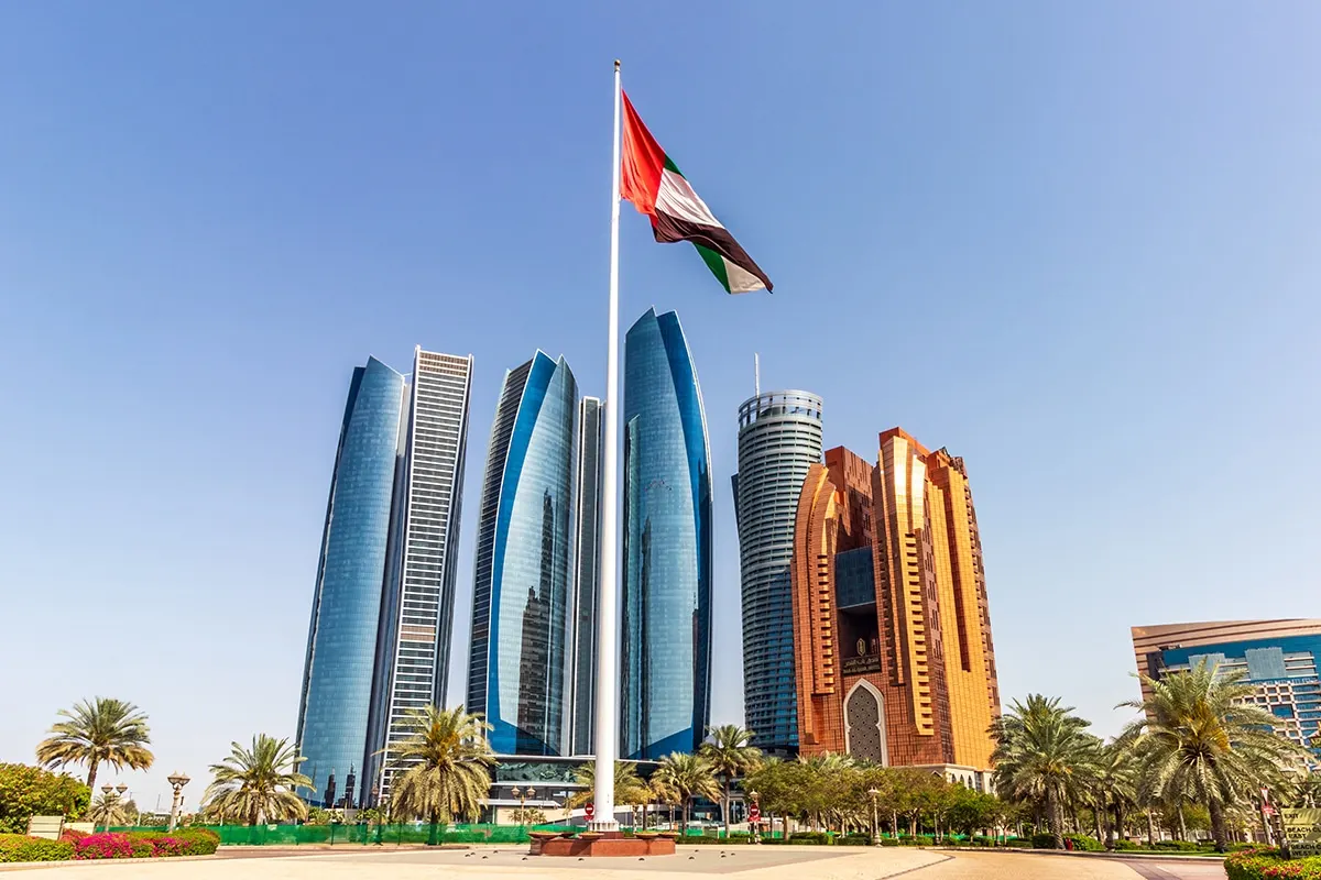 The UAE flag flying on a tall flagpole in Abu Dhabi in front of the Etihad Towers and other high-rise buildings, with palm trees under a clear blue sky.