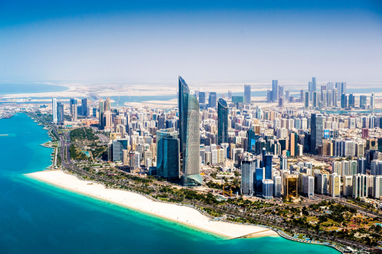 Aerial view of the Abu Dhabi skyline, UAE, with high-rise towers along a white sand beach and turquoise waters.