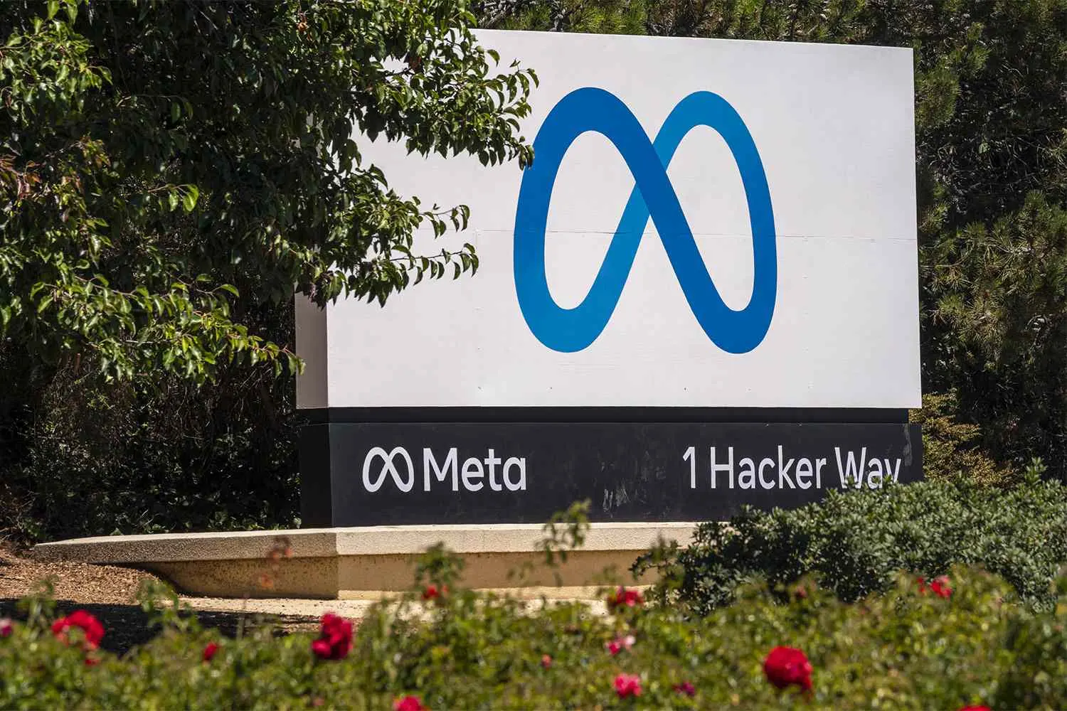 The Meta infinity logo and name on the sign at 1 Hacker Way, the company's headquarters address, behind red flowers