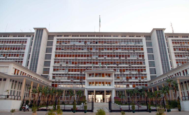 The Government Palace in Algiers, a white multi-storey building with orange window shades and the Algerian flag on its roof, seen from the gated main entrance lined with palm trees