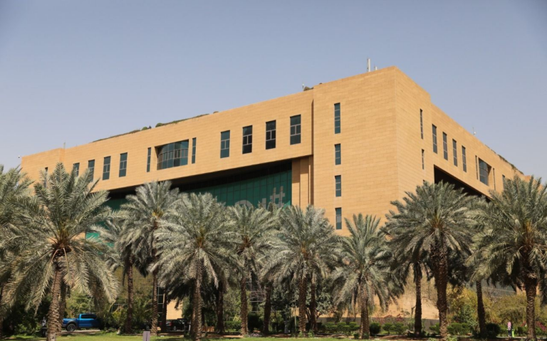 A sand-coloured building of the Saudi Ministry of Municipalities and Housing with a green glass entrance, behind a row of date palms under a clear sky